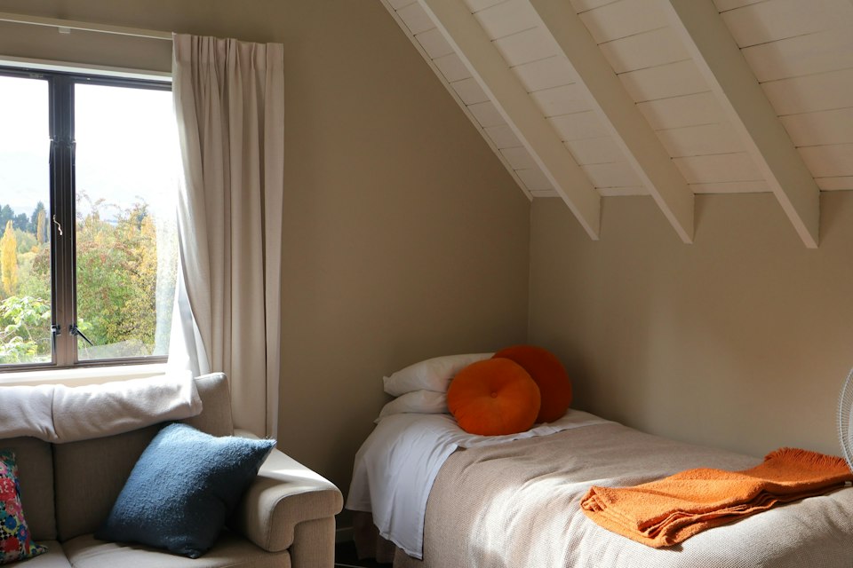 Bedroom with a sloped beamed ceiling, a single bed with orange pillows, a sofa and a window onto trees