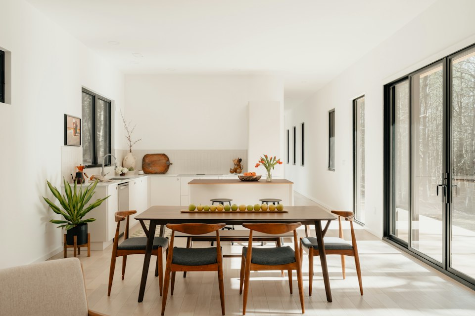 Dining area with a wooden table and chairs, the kitchen beyond, and tall glass doors to the right