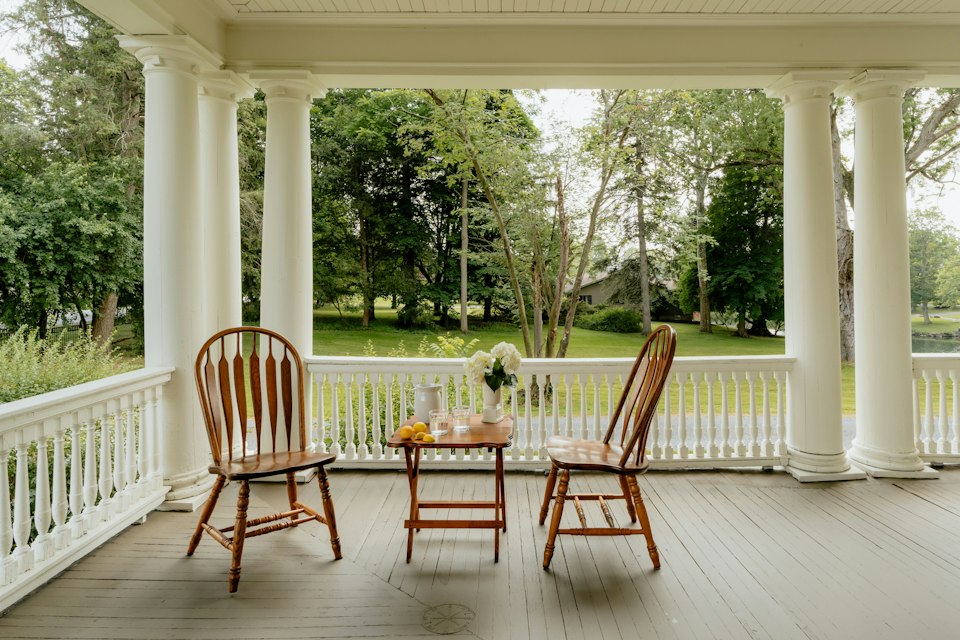 Covered porch with white columns and railings, two wooden chairs and a small table, looking out over trees and lawn