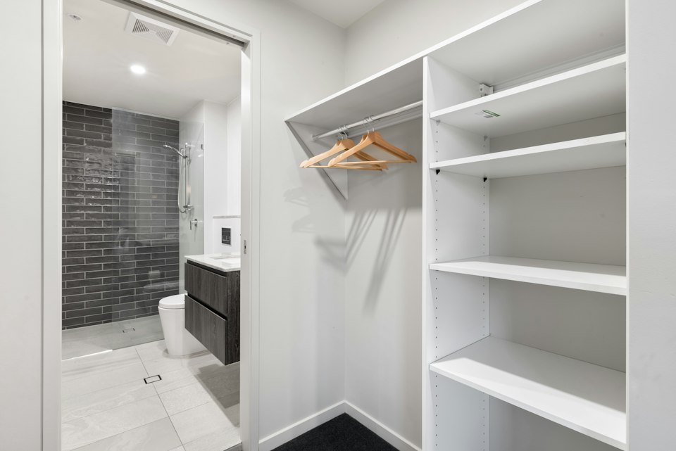 Closet with white open shelving and a hanging rail, with a tiled bathroom visible through the doorway